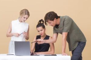 Three people are looking at a laptop while one of them is holding a pen.